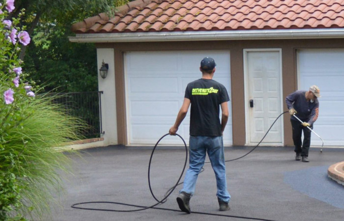 Two individuals on a residential driveway, one holding a pressure washing hose aimed at the ground and the other standing with a coiled hose.
