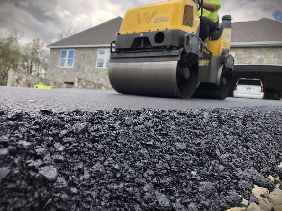 A close-up of fresh black asphalt with a steamroller and worker in the background, in front of a stone facade building under a cloudy sky.