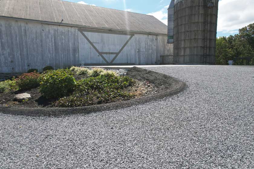 A gravel area with a garden bed, a large barn with a sliding door, and a metal silo under a partly cloudy sky.